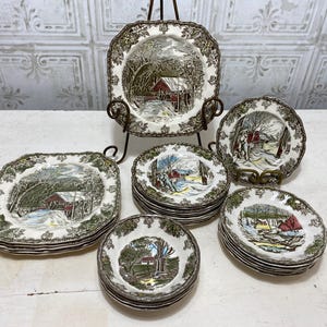 May include: A set of 10 vintage ceramic plates featuring a winter scene with a red barn and a bridge. The plates are in various sizes and are stacked on top of each other.