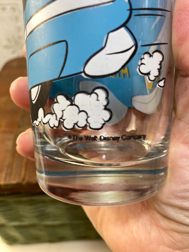 May include: A clear glass drinking glass with a blue and white cartoon image of a car with smoke coming out of the exhaust. The text "The Walt Disney Company" is printed on the bottom of the glass.