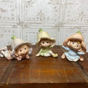 May include: Three ceramic figurines of elves wearing flower-shaped hats. The elf on the left is lying down with a pink and white hat. The elf in the middle is sitting up with a green and white hat. The elf on the right is sitting up with a blue and white hat.