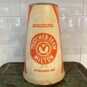 May include: A vintage, white, cylindrical container with orange accents. The container has a red and white logo that reads "Thatcher Farm Milton" and "Established 1891". The text "One Half Pint Liquid Penalty for Re-use Twenty-five Dollars" is printed on the container.
