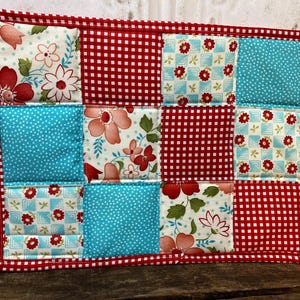 May include: A rectangular quilted potholder featuring a patchwork design of twelve squares. The fabric patterns include red and white gingham, turquoise with white dots, and a floral print with red and pink flowers on a white background.