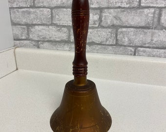 Puede incluir: Una campana de escuela de latón vintage con un mango de madera. La campana tiene una pátina desgastada y envejecida y el mango es de color marrón oscuro con veta visible.
