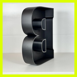 May include: Black plastic letter B, standing upright on a white surface. The letter is hollow and has a three-dimensional design.