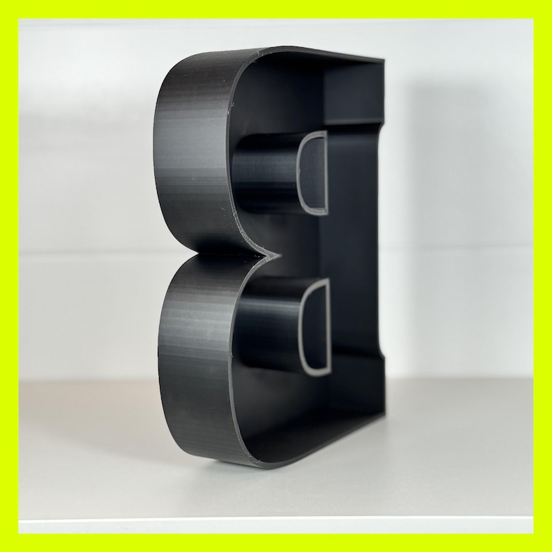 May include: Black plastic letter B, standing upright on a white surface. The letter is hollow and has a three-dimensional design.