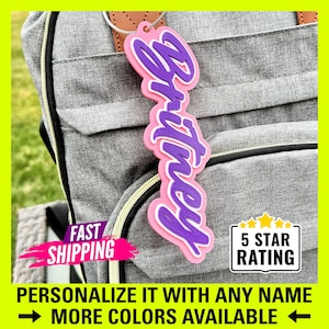 May include: A pink and purple personalized name tag with the name "Britney" on it. The tag is attached to a gray backpack with black straps. The tag is a great way to personalize your belongings.