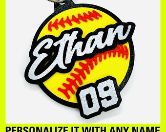 Könnte beinhalten: Ein gelber Softball-Anhänger mit roten Nähten und schwarzem Rand. Der Name "Ethan" ist in Weiß mit schwarzem Rand auf dem Softball geschrieben. Die Nummer "09" ist in Weiß mit schwarzem Rand auf der Unterseite des Anhängers geschrieben.