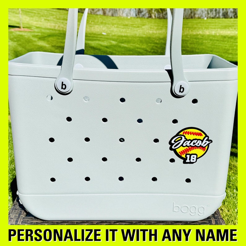May include: Grey tote bag with a softball design and the name "Jacob" and the number "18" on it. The bag has a white "b" on each handle.