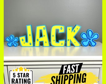 May include: Yellow and blue 3D letters spelling the name "JACK" with a blue and yellow flower on each side. The letters are decorated with blue dots. The letters are sitting on a white surface.