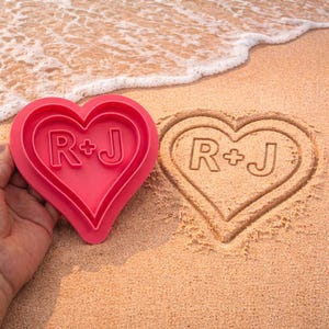 May include: A pink heart-shaped mold with the initials "R+J" is held next to a heart-shaped imprint in the sand, also with the initials "R+J". The scene is set on a sandy beach with gentle waves in the background.