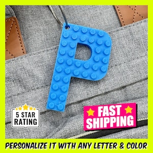 May include: Blue plastic letter P keychain shaped like a Lego brick.  The keychain is hanging on a gray fabric background.  The text "FAST SHIPPING" is visible in the image.