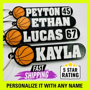 May include: Four black and white personalized basketball keychain tags with a basketball graphic. The tags read "Peyton 45", "Ethan", "Lucas 67", and "Kayla".