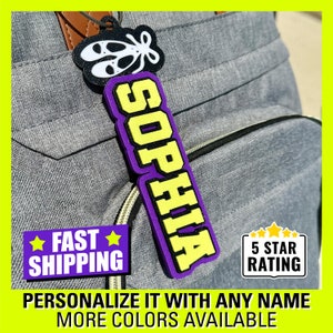 May include: A purple and yellow personalized name tag with the name "SOPHIA" on it. The tag is attached to a gray backpack. The tag is available in multiple colors.