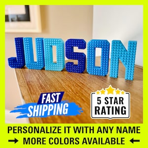 May include: Blue and white plastic letters spelling the name "JUDSON" in a Lego-style design. The letters are arranged on a wooden surface. The text "FAST SHIPPING" and "5 STAR RATING" are visible below the letters.