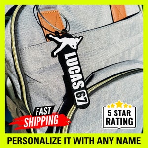 May include: A white baseball player silhouette on a black luggage tag with the name "Lucas 67" printed in white. The tag is attached to a gray backpack with a brown leather strap. The text "FAST SHIPPING" is in red and the text "5 STAR RATING" is in yellow.