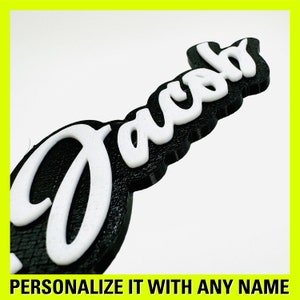 May include: A black and white 3D printed name sign with the name "Jacob" in a cursive font. The sign is made of a textured material and has a raised edge.
