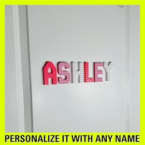 May include: A white door with the name "ASHLEY" spelled out in pink and white Lego bricks. The text "PERSONALIZE IT WITH ANY NAME" is at the bottom of the image.