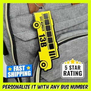 May include: A yellow school bus-shaped luggage tag with a black outline and the text "BUS NUMBER 13B". The tag is attached to a gray backpack with a zipper. The text "FAST SHIPPING" and "5 STAR RATING" are also visible in the image.