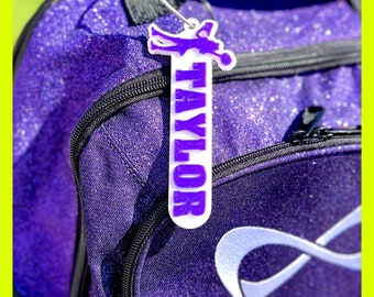 Puede incluir: Mochila de purpurina morada con una etiqueta blanca que dice "TAYLOR" en letras de purpurina morada. La etiqueta está unida a la mochila con una cuerda plateada.