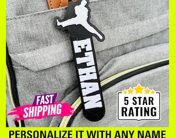 May include: Black and white personalized luggage tag with a basketball player silhouette and the name "ETHAN" printed vertically. The tag is attached to a gray backpack with a brown leather strap. The text "5 STAR RATING" is visible in a white rectangle with yellow stars.