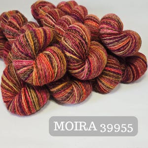 Pode incluir: Uma coleção de novelos de lã coloridos em tons de vermelho, laranja, amarelo e castanho. A lã é bem enrolada e parece macia. O texto "MOIRA 39955" é exibido em um retângulo cinza na parte inferior. Ideal para projetos de tricô e crochê.