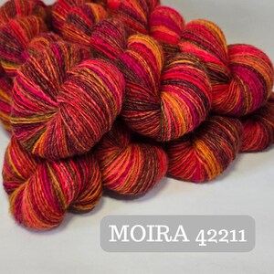 Pode incluir: Uma coleção de novelos de fios vibrantes em tons de vermelho, laranja e marrom. O fio é enrolado firmemente, exibindo um padrão listrado. O texto "MOIRA 42211" é exibido em um retângulo cinza.