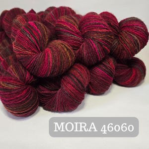 Pode incluir: Close-up de vários novelos de fio em tons variados de vermelho, rosa e castanho. O fio é enrolado com firmeza, com variações visíveis de textura e cor. O texto "MOIRA 4606o" é exibido em um retângulo cinza na parte inferior.