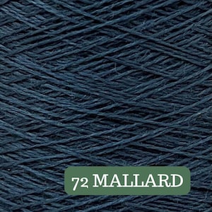 May include: Close-up of a ball of dark blue yarn. The yarn is tightly wound, with visible strands creating a textured surface. A green rectangular label at the bottom reads "72 MALLARD" in white text.