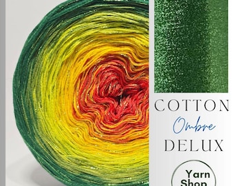 May include: A vibrant ball of Cotton Ombre Deluxe yarn, featuring a gradient of colors from green to yellow to red. The yarn has a metallic sheen. The image also includes the text "Cotton Ombre Deluxe" and a logo for "Yarn Shop by Stay Alive."