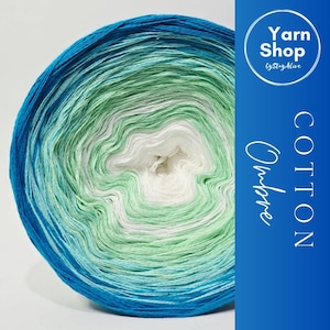 May include: A ball of ombre cotton yarn in shades of blue, green, and white. The yarn is labelled "Cotton Ombre" and "Yarn Shop by StayAlive". The colours transition smoothly, creating a gradient effect, ideal for knitting and crochet projects.