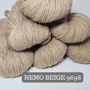 Pode incluir: Um monte de novelos de lã bege. A lã é de cor bege claro, com um aspeto ligeiramente texturizado. O texto "NEMO BEIGE 9698" é exibido num retângulo cinzento na parte inferior da imagem. Ideal para projetos de tricô e crochê.