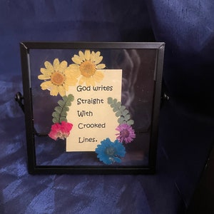 Peut inclure: Une image encadrée noire avec une carte blanche à l'intérieur qui dit "Dieu écrit droit avec des lignes courbes." La carte est entourée de fleurs pressées dans des tons de jaune, rose, bleu et vert.