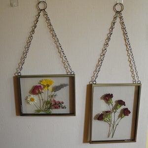 Puede incluir: Dos arreglos de flores prensadas enmarcados colgados en una pared blanca. El marco superior presenta una margarita amarilla, una rosa rosa y una flor morada. El marco inferior presenta un ramo de claveles rojos.