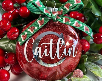 May include: A red glitter Christmas ornament with the name "Caitlin" in white script. The ornament has a silver metal top and is adorned with a green ribbon patterned with Santa hats. Red berries and green leaves surround the ornament.