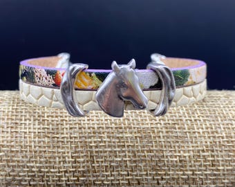 Op de afbeelding: Een armband met twee lagen, versierd met een zilveren paardenkop. De bovenste laag heeft een bloemenpatroon met paarse randen, terwijl de onderste laag een gestructureerd, gebroken wit ontwerp heeft. De armband wordt gepresenteerd op een gestructureerd, bruin oppervlak.