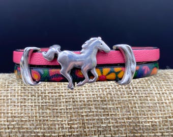 Op de afbeelding: Een gestapelde leren armband met een zilveren paardenbedel. De bovenste laag is felroze en de onderste laag heeft een bloemenpatroon met geel, oranje en groen. De armband heeft zilveren accenten aan elk uiteinde.