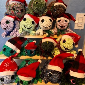 May include: A display of crocheted turtle ornaments, each wearing a Santa hat. The turtles are in shades of green, blue, and brown, arranged on wooden shelves. The hats have white trim, with some featuring snowflake patterns.