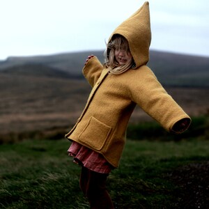 May include: A young person wearing a yellow hooded jacket with a brown skirt and brown leggings. The person is standing in a field of green grass.