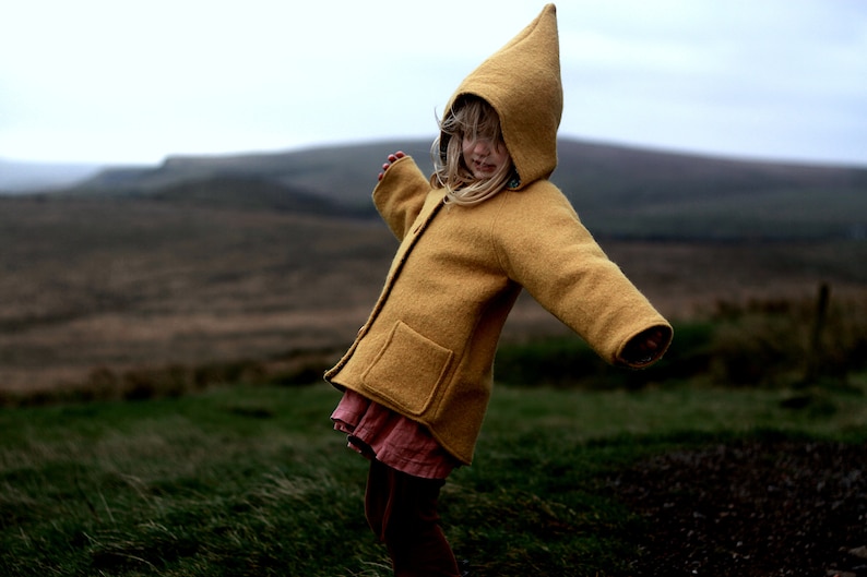 May include: A young person wearing a yellow hooded jacket with a brown skirt and brown leggings. The person is standing in a field of green grass.