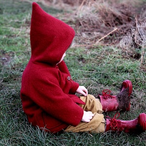 May include: A child wearing a red hooded jacket sits in a grassy field. The child is wearing red rain boots and has their back to the camera.