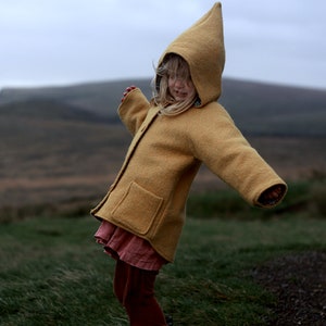 May include: A young girl wearing a yellow hooded jacket with a pointed hood, standing in a field with a blurry background.