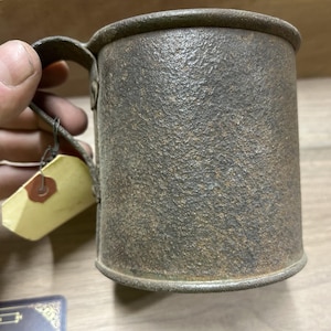 May include: A cylindrical metal mug with a handle, showing signs of age and wear. The mug has a textured, dark brown surface and a simple, utilitarian design. A small tag is attached to the handle.