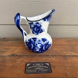 May include: A white ceramic pitcher with a blue floral design. The pitcher has a curved handle and a spout. The blue floral pattern covers the body of the pitcher. The pitcher is sitting on a wooden surface.