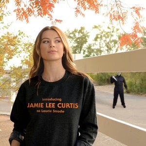 May include: A woman wearing a dark gray sweatshirt with the text "Introducing Jamie Lee Curtis as Laurie Strode" in orange. She is standing in front of a white picket fence with a figure in a black jumpsuit and a mask holding a knife in the background.