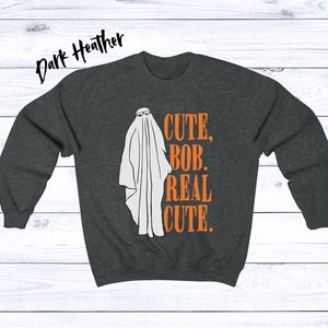Puede incluir: Sudadera gris oscuro con una ilustraci&oacute;n de fantasma blanco con gafas de sol. El texto naranja dice "CUTE, BOB, REAL CUTE". Un dise&ntilde;o divertido y espeluznante para Halloween.