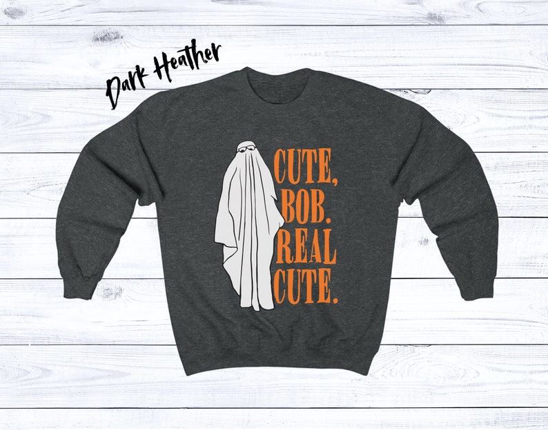 Puede incluir: Sudadera gris oscuro con una ilustraci&oacute;n de fantasma blanco con gafas de sol. El texto naranja dice "CUTE, BOB, REAL CUTE". Un dise&ntilde;o divertido y espeluznante para Halloween.