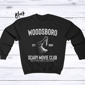 May include: Black sweatshirt with a white graphic design. The graphic features a hand holding a knife and the text "Woodsboro Est 1996 Scary Movie Club What's your favorite scary movie?"