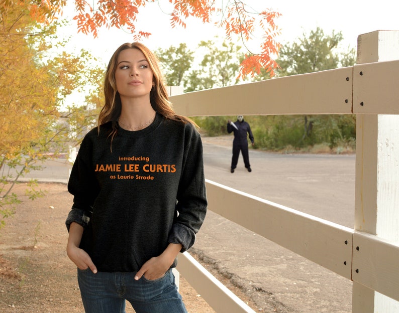 May include: A woman wearing a dark gray sweatshirt with the text "Introducing Jamie Lee Curtis as Laurie Strode" in orange. She is standing in front of a white picket fence with a figure in a black jumpsuit and a mask holding a knife in the background.