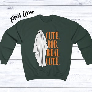 Puede incluir: Sudadera verde bosque con un gr&aacute;fico de fantasma blanco con gafas de sol. El texto "CUTE, BOB, REAL CUTE." est&aacute; impreso en naranja. Una divertida prenda de vestir con tem&aacute;tica de Halloween.