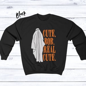 Puede incluir: Sudadera negra con un fantasma blanco con gafas de sol. El texto naranja dice "CUTE, BOB, REAL CUTE". La palabra "Black" est&aacute; escrita en la esquina superior izquierda.