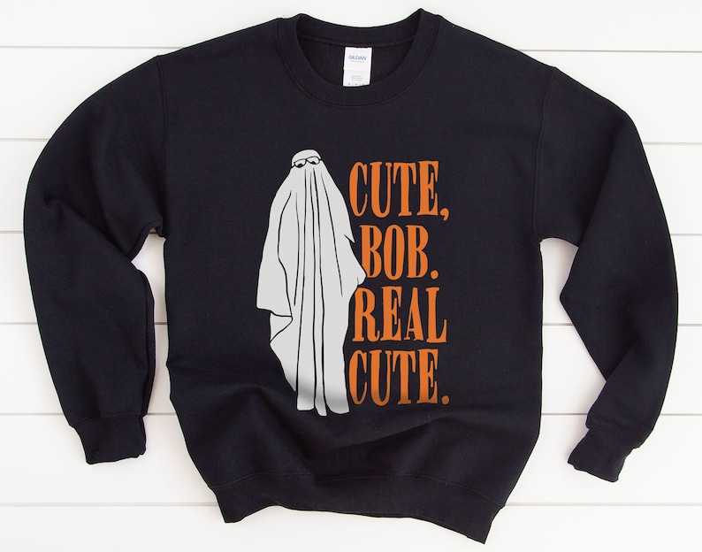 Puede incluir: Sudadera negra con un fantasma blanco con gafas. El texto "CUTE, BOB, REAL CUTE" est&aacute; impreso en naranja. Una prenda con tem&aacute;tica de Halloween.